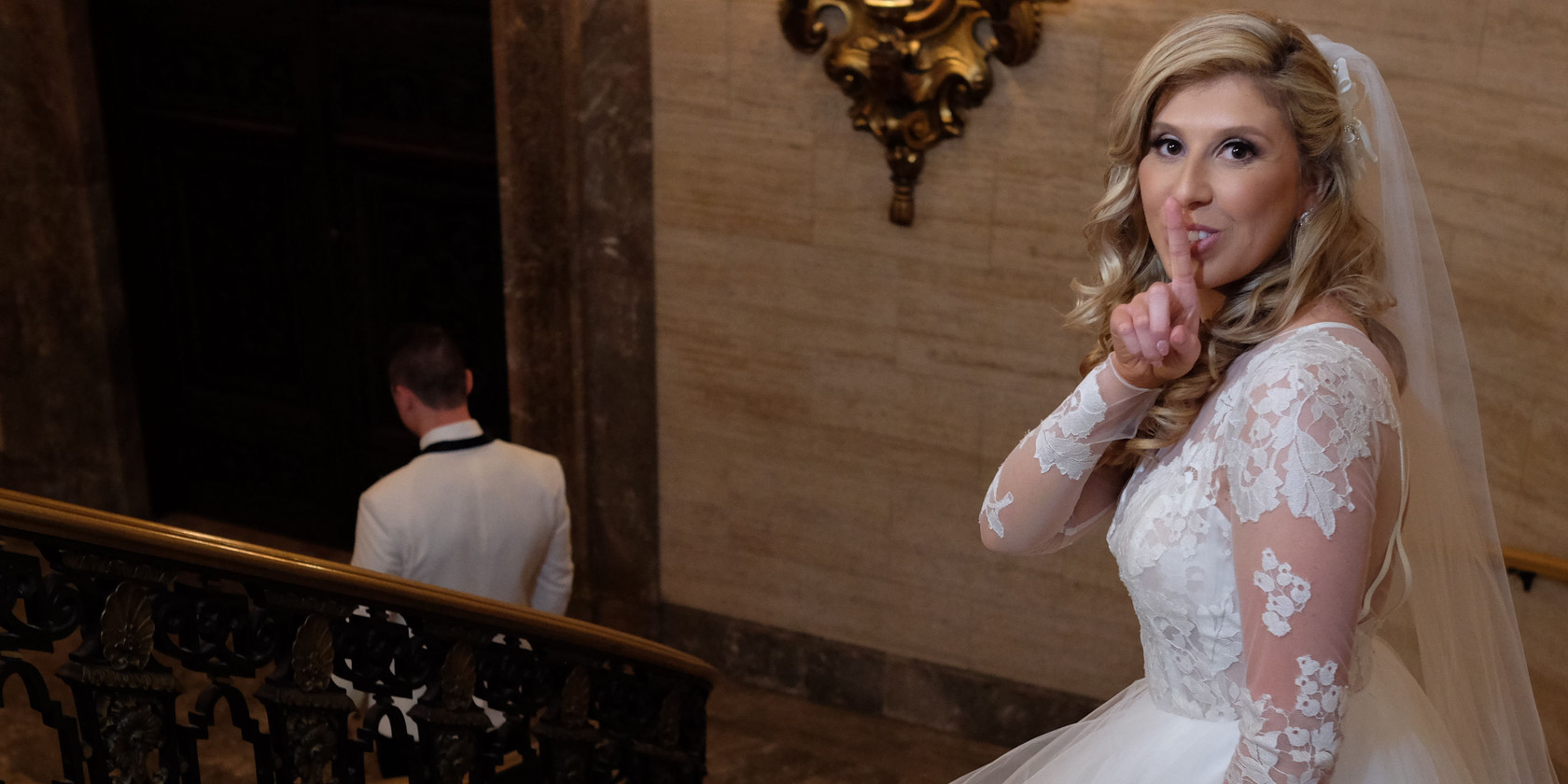 A bride shares a quiet playful gesture while the groom descends an ornate staircase